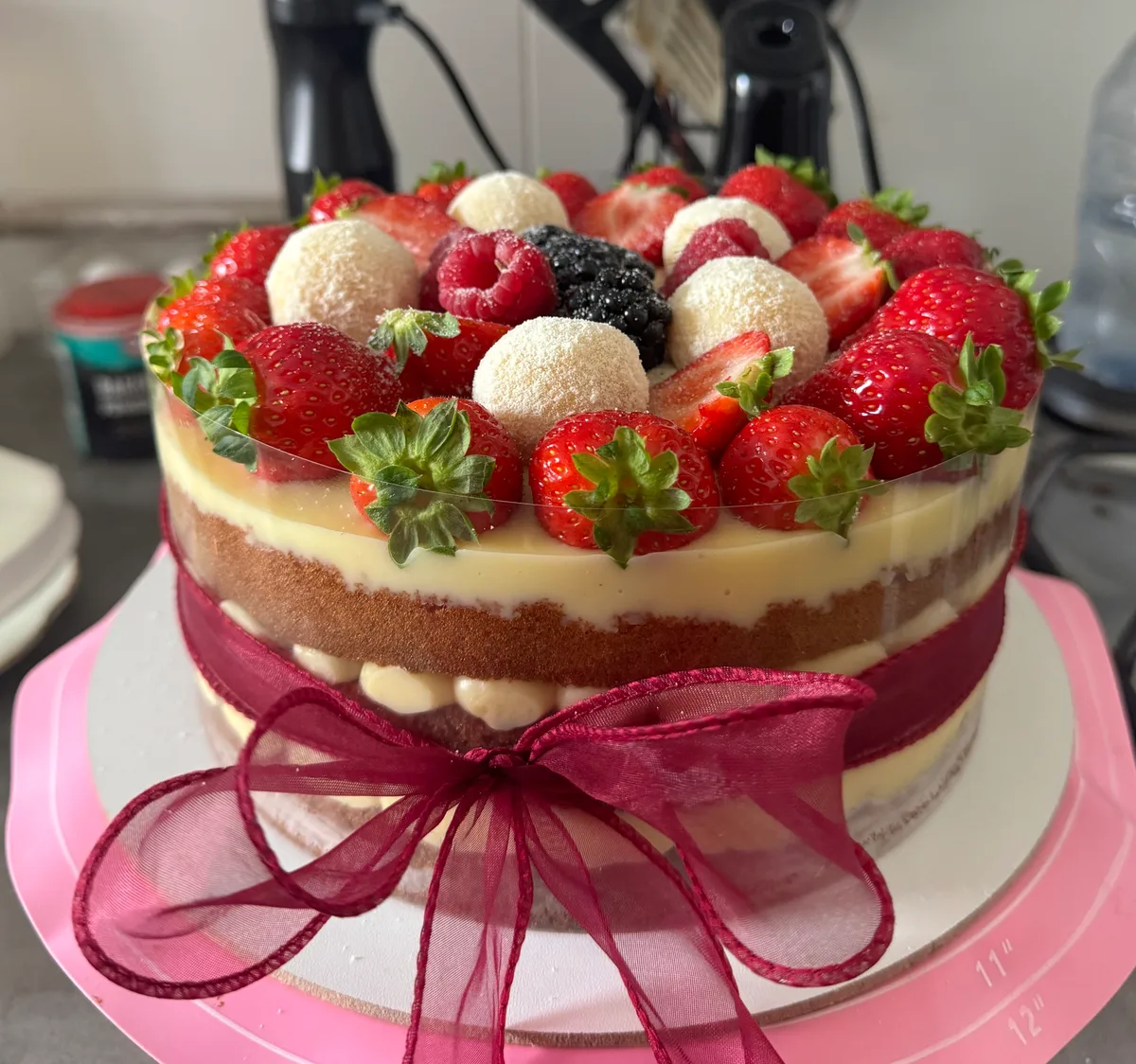 Naked cake com camadas de creme aparentes, morangos frescos, frutas vermelhas e trufas brancas no topo e um laço bordô